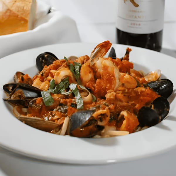 Seafood pasta with mussels, shrimp, and clams in red sauce, with wine and bread in the background.